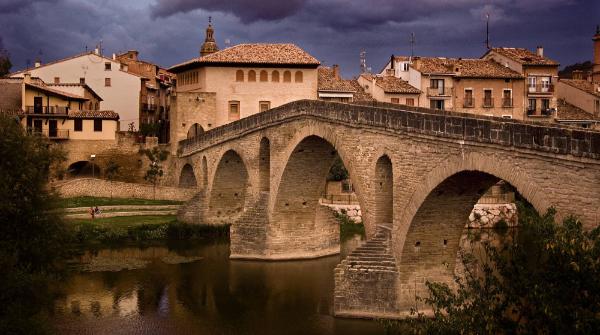 Image du coucher du soleil dans laquelle le pont roman de Puente la Reina est vu au premier plan et la ville derrière